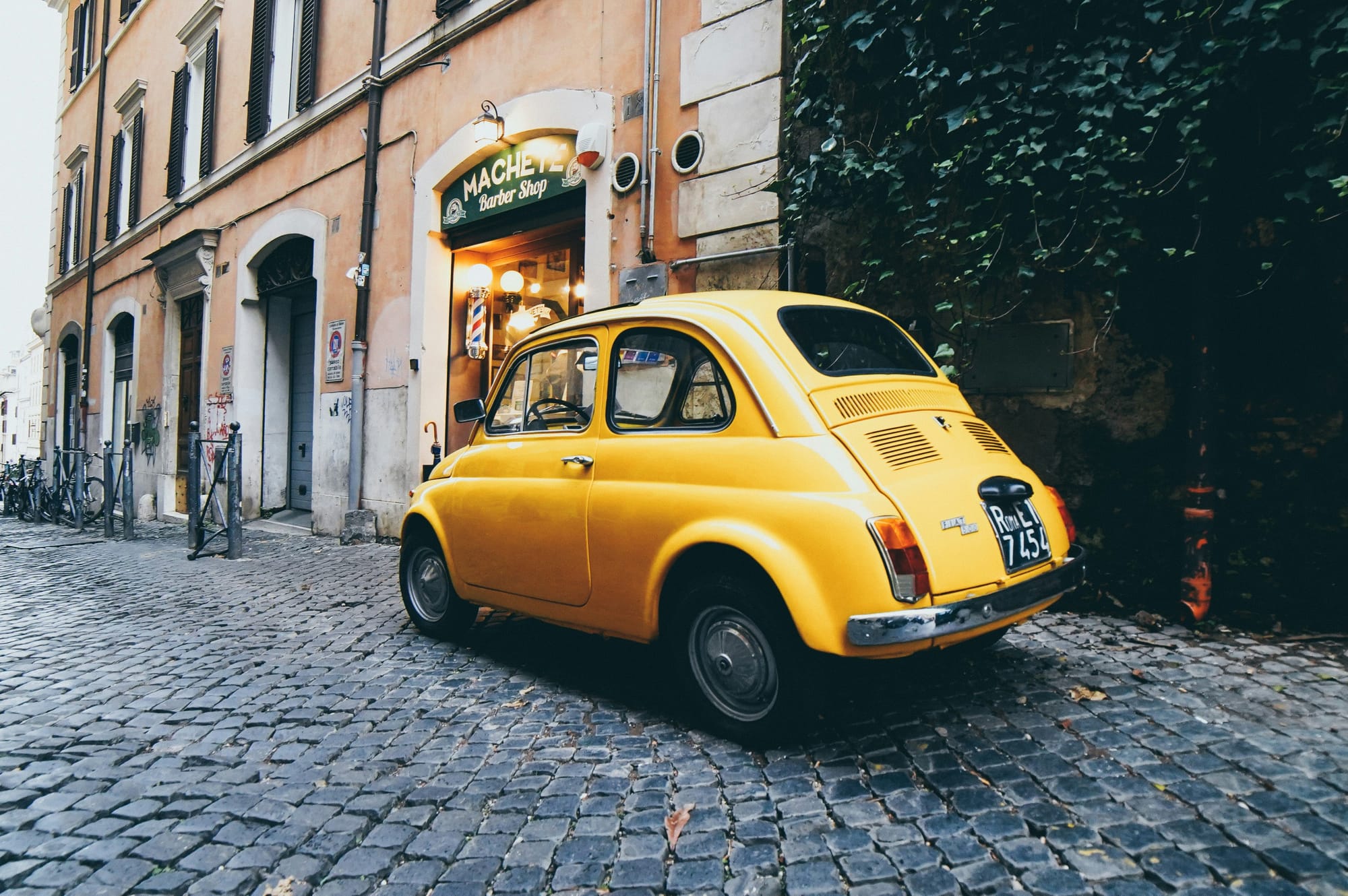 A narrow cobblestone street in Trastevere, Rome, with ivy-covered buildings, a traditional trattoria and a classic iconic Italian car.